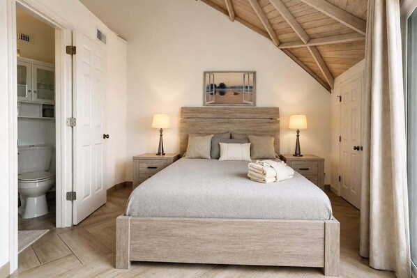 A bedroom with a wooden headboard, a bed with a grey bedspread, two bedside tables with lamps, and a view of a lake through an open window. - ROOM 04 - Lake Sumner (Clermont)