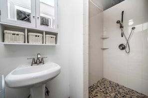 Une salle de bain avec un lavabo blanc, des paniers en osier sur des étagères et une douche avec un sol carrelé.