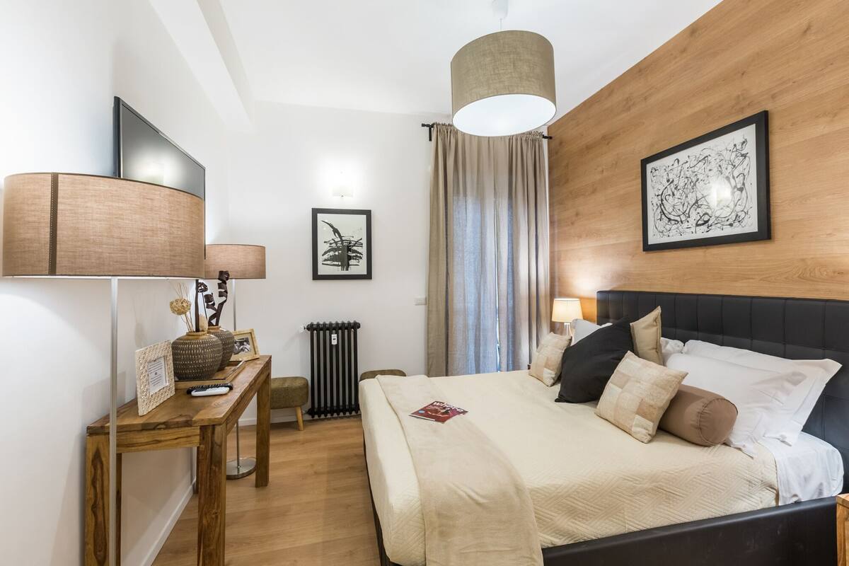 A hotel room with a bed, bedside table, lamp, artwork, and a radiator.
