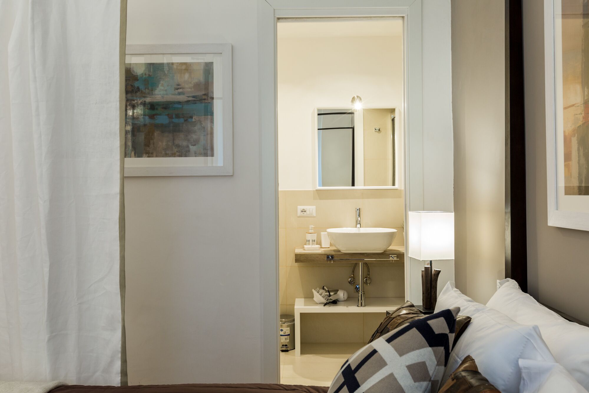 Habitación de hotel con una cama, una mesita de noche, una lámpara, un espejo, un lavamanos y un cuadro en la pared.