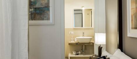 Habitación de hotel con una cama, una mesita de noche, una lámpara, un espejo, un lavamanos y un cuadro en la pared.