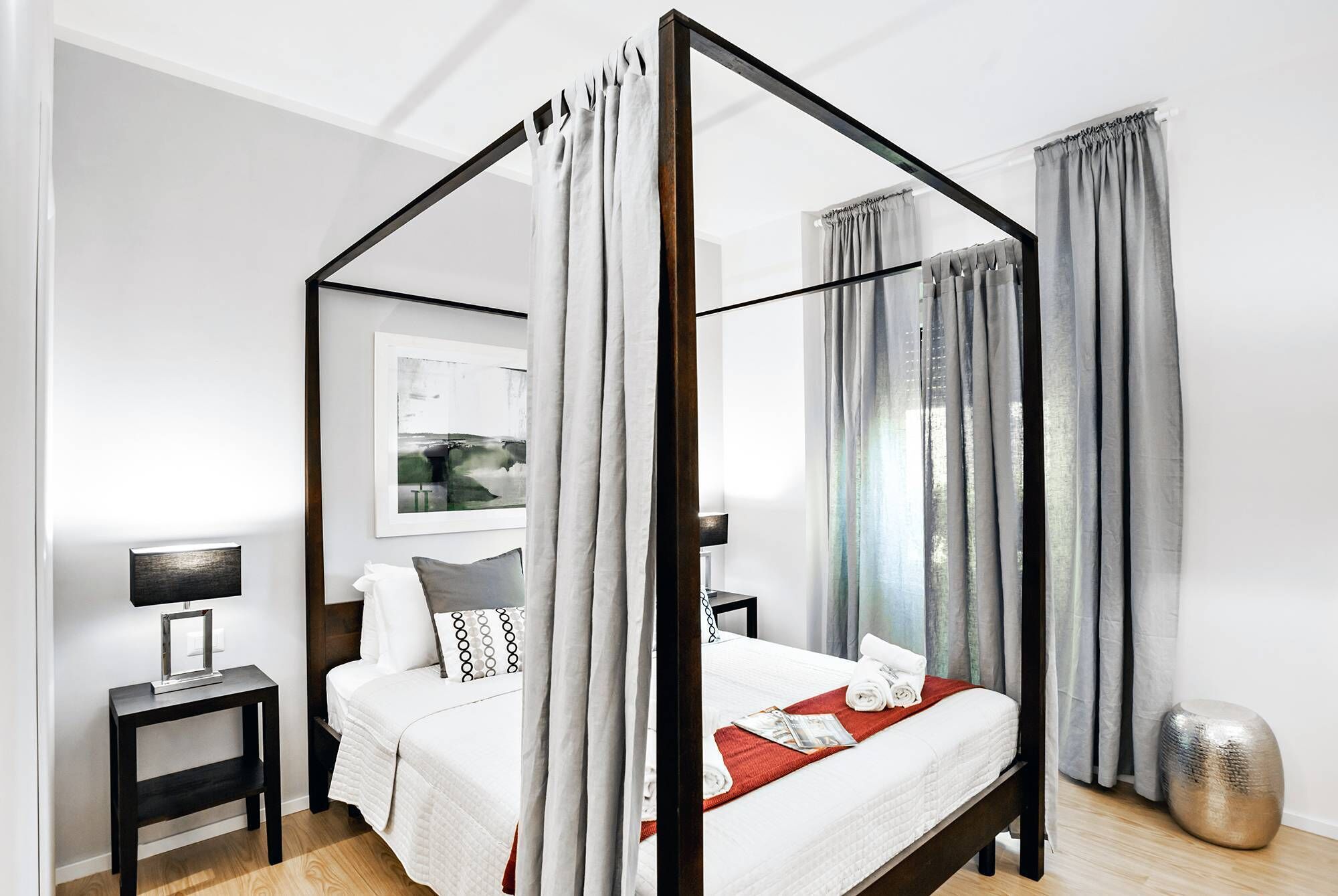 A four-poster bed with a white bedspread and red throw, a bedside table with a lamp, and a framed picture on the wall.