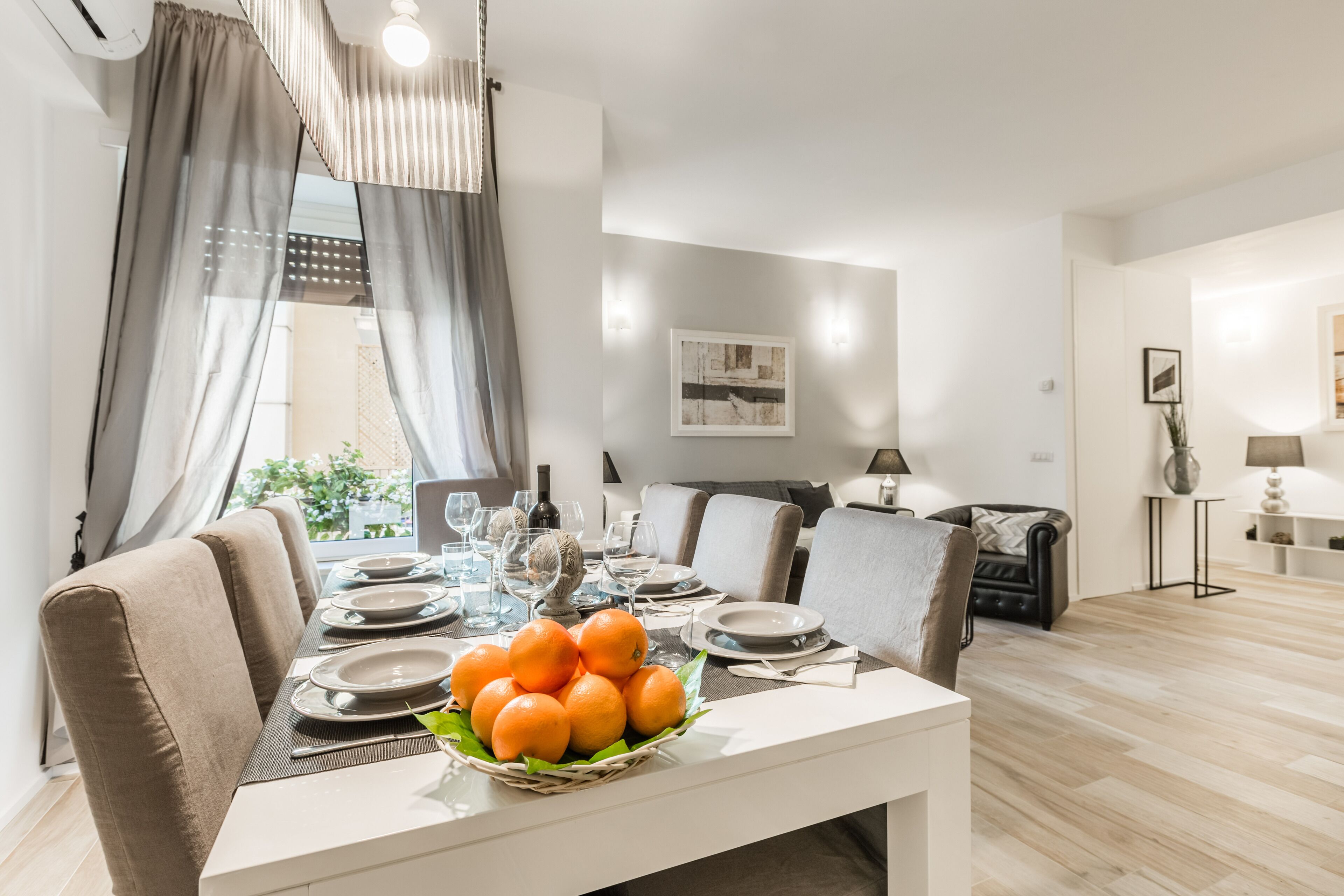A modern dining area with a table set for four, a basket of oranges, and a sofa in the background.