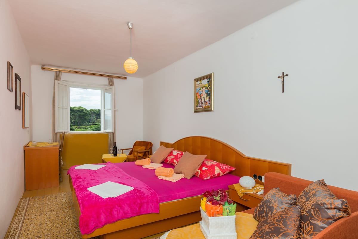 A bedroom with a bed, a wooden nightstand, a bedside table, a window, and a cross on the wall.