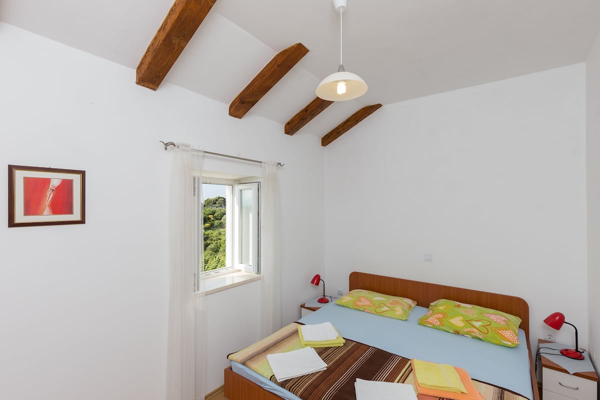 A bedroom with a wooden ceiling, a bed with colorful bedding, a window with white curtains, and a red lamp on the nightstand.