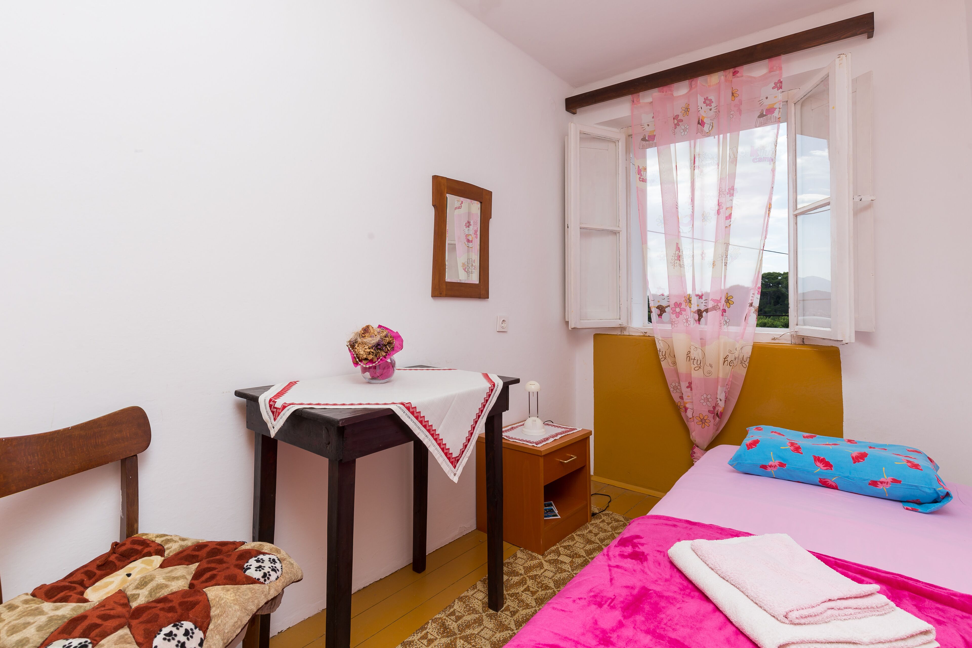 A bedroom with a bed, a bedside table, a chair, a window with curtains, and a small table with a vase.