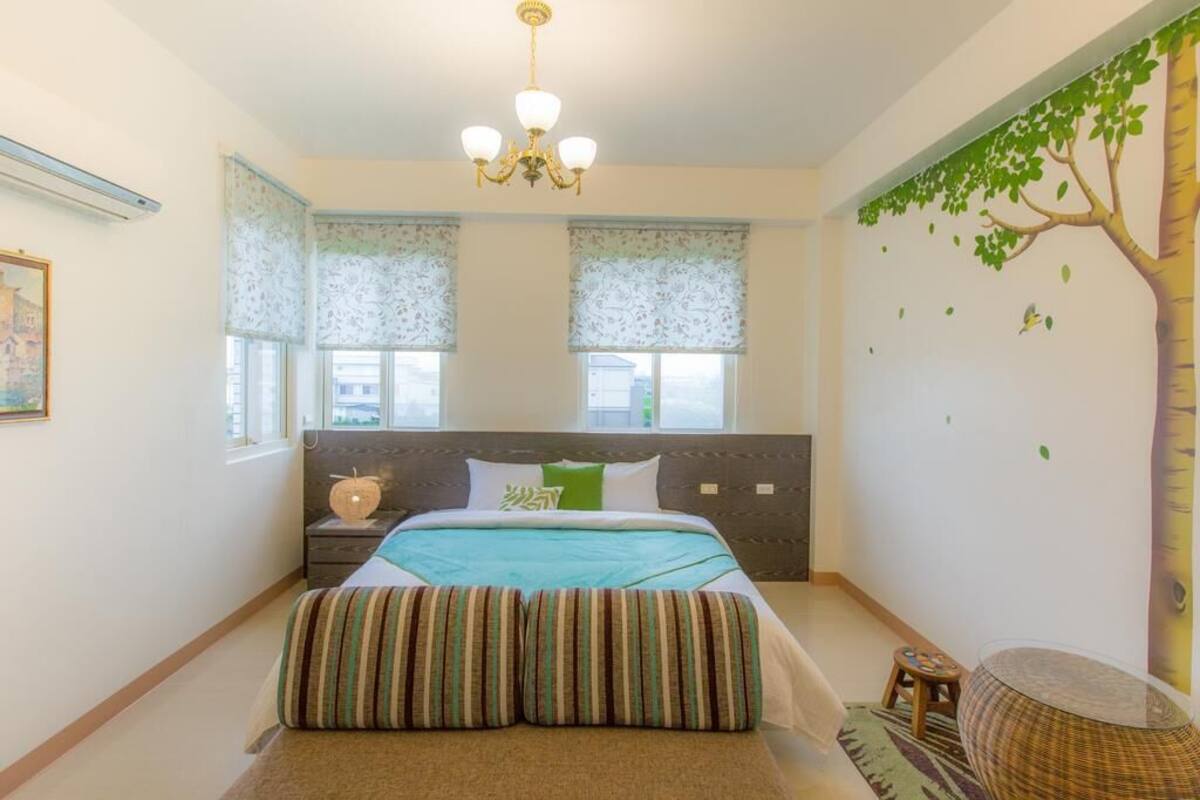 A bedroom with a bed, a nightstand, a chair, a window with a view, and a wall with a tree mural.