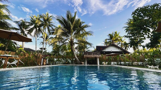 A swimming pool with clear blue water, surrounded by palm trees and a thatched-roof structure. - The Village Siargao (General Luna)