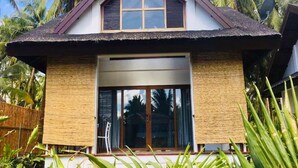 A two-story house with a thatched roof, large windows, and a sliding glass door. - The Village Siargao (General Luna)