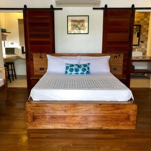 A wooden bed with a white mattress and a blue and white pillow, a framed picture on the wall, and a kitchen area in the background. - The Village Siargao (General Luna)
