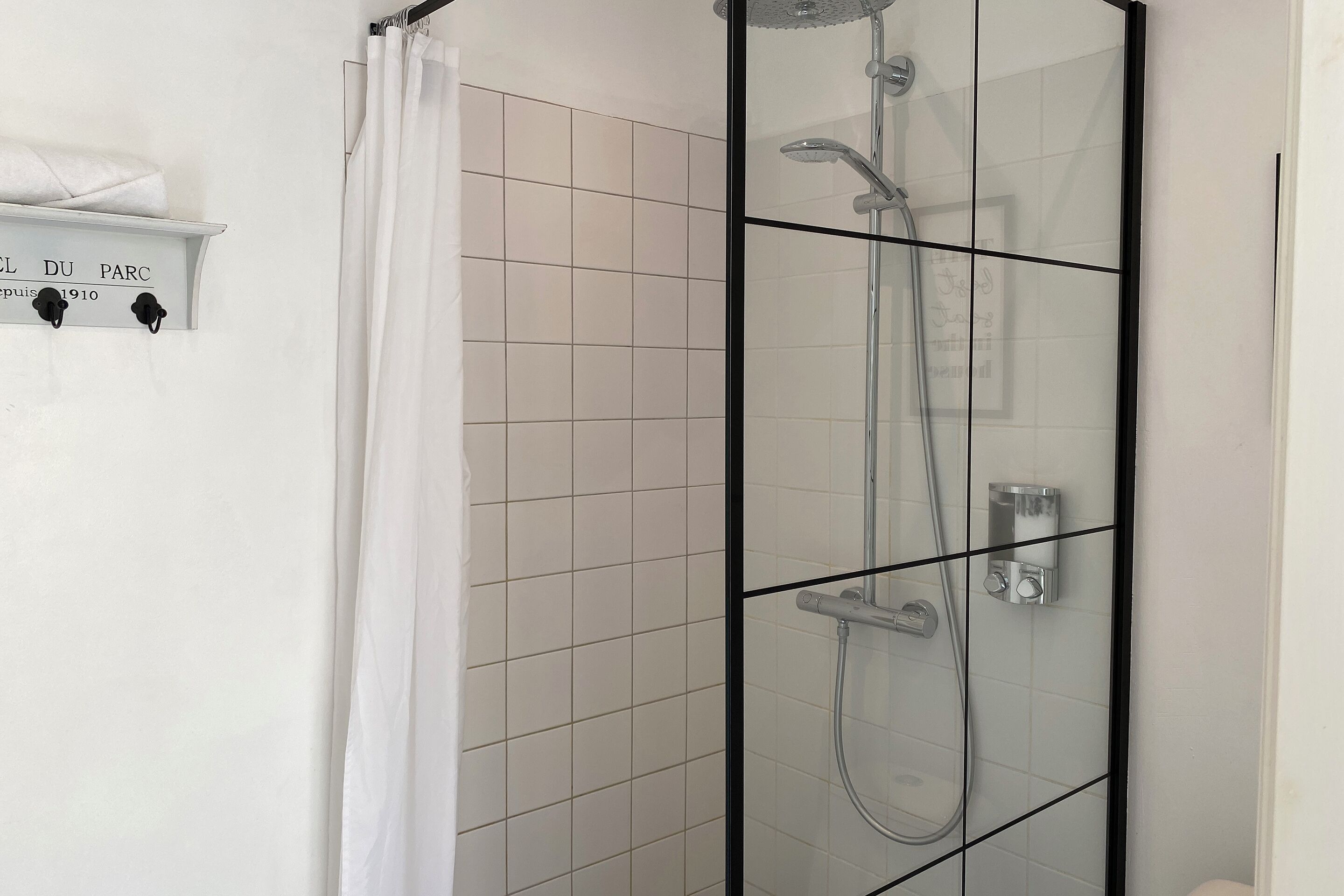 A glass-enclosed shower with a white tiled wall, a white shower curtain, and a shelf with a towel.