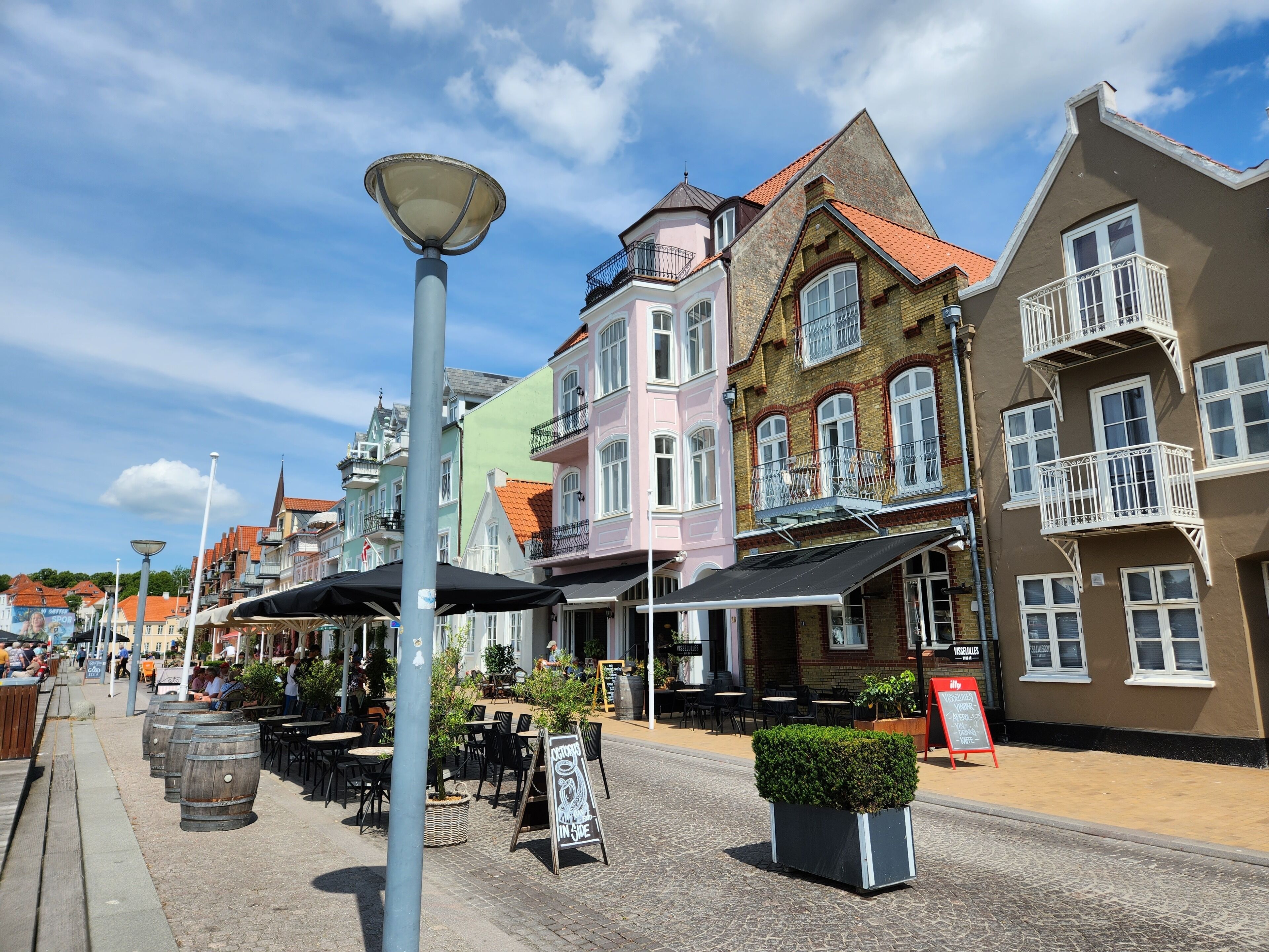 A cobblestone street lined with colorful buildings, outdoor seating, and a signboard.