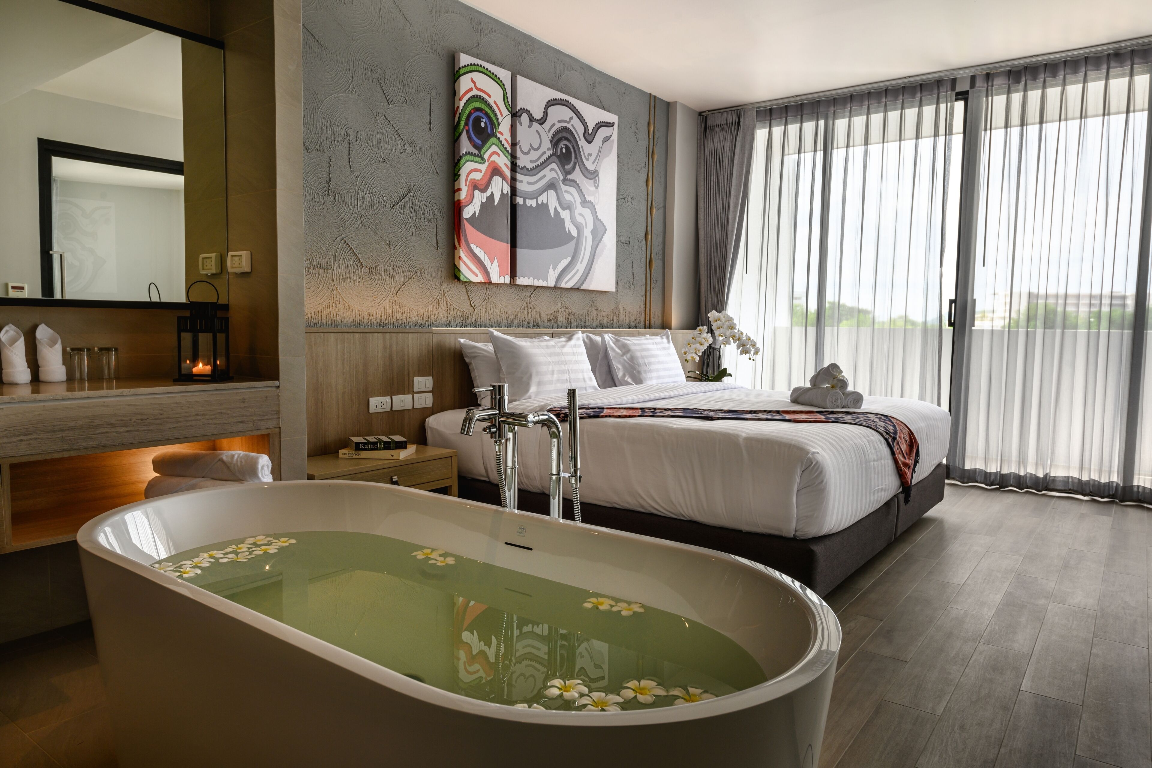 A modern hotel room with a large bathtub, a bed with white linens, a large mirror, and artwork on the wall.