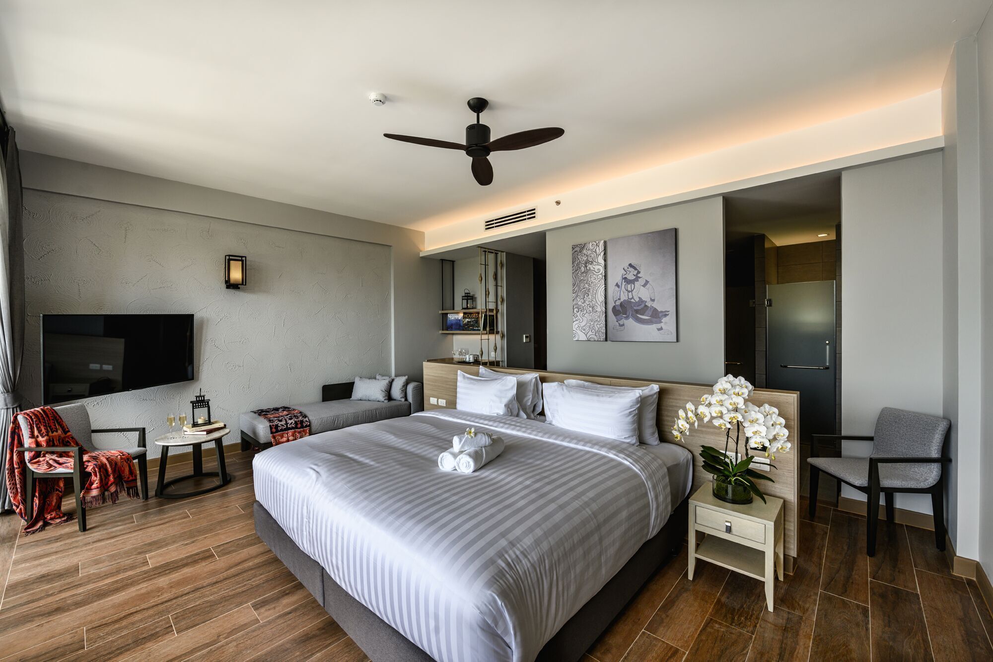 A modern hotel room with a large bed, a ceiling fan, a flat-screen TV, a small table with a lamp, and a chair.