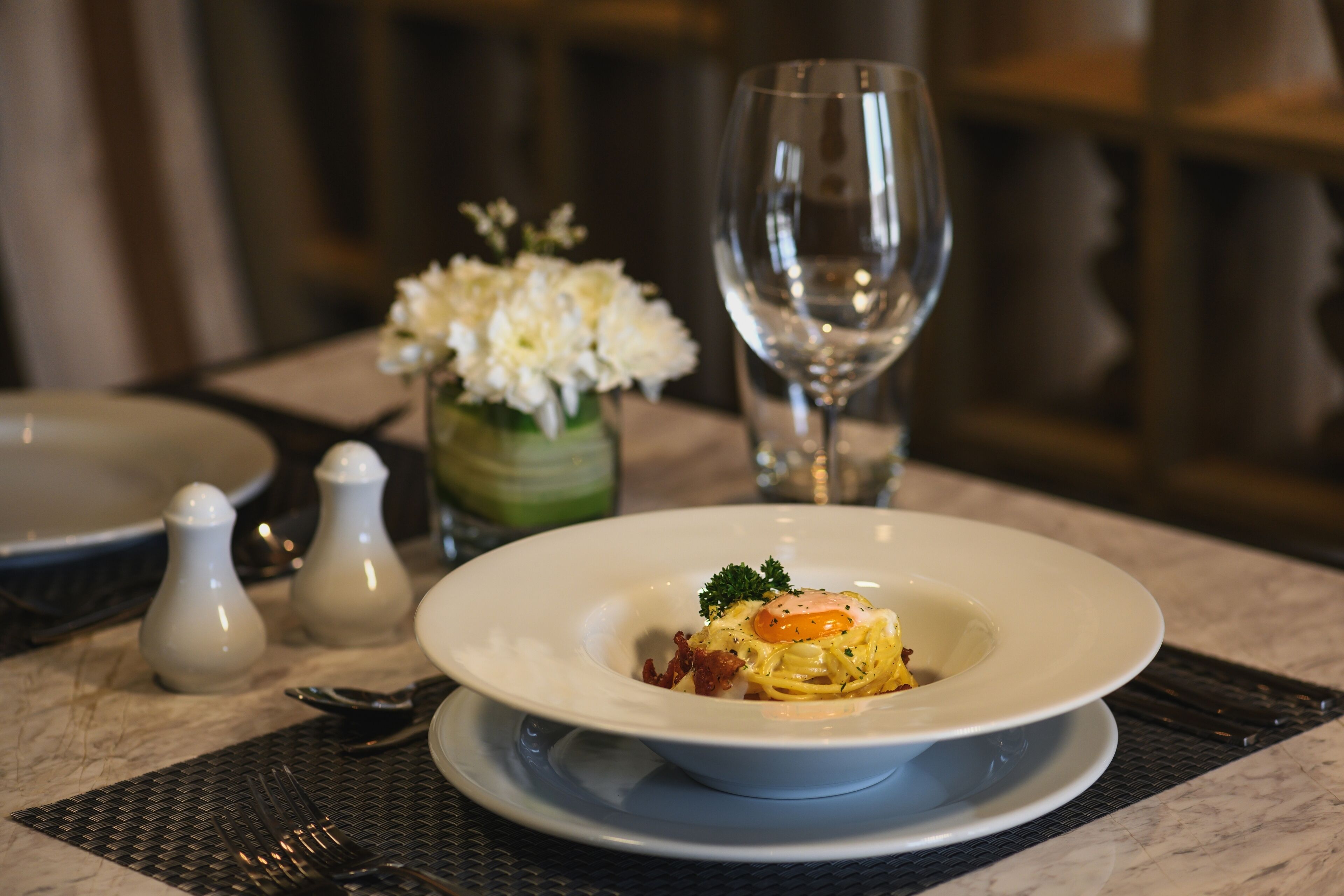 Une table à manger avec une assiette de pâtes, un verre d’eau, des salières et des poivrières, et un vase de fleurs.