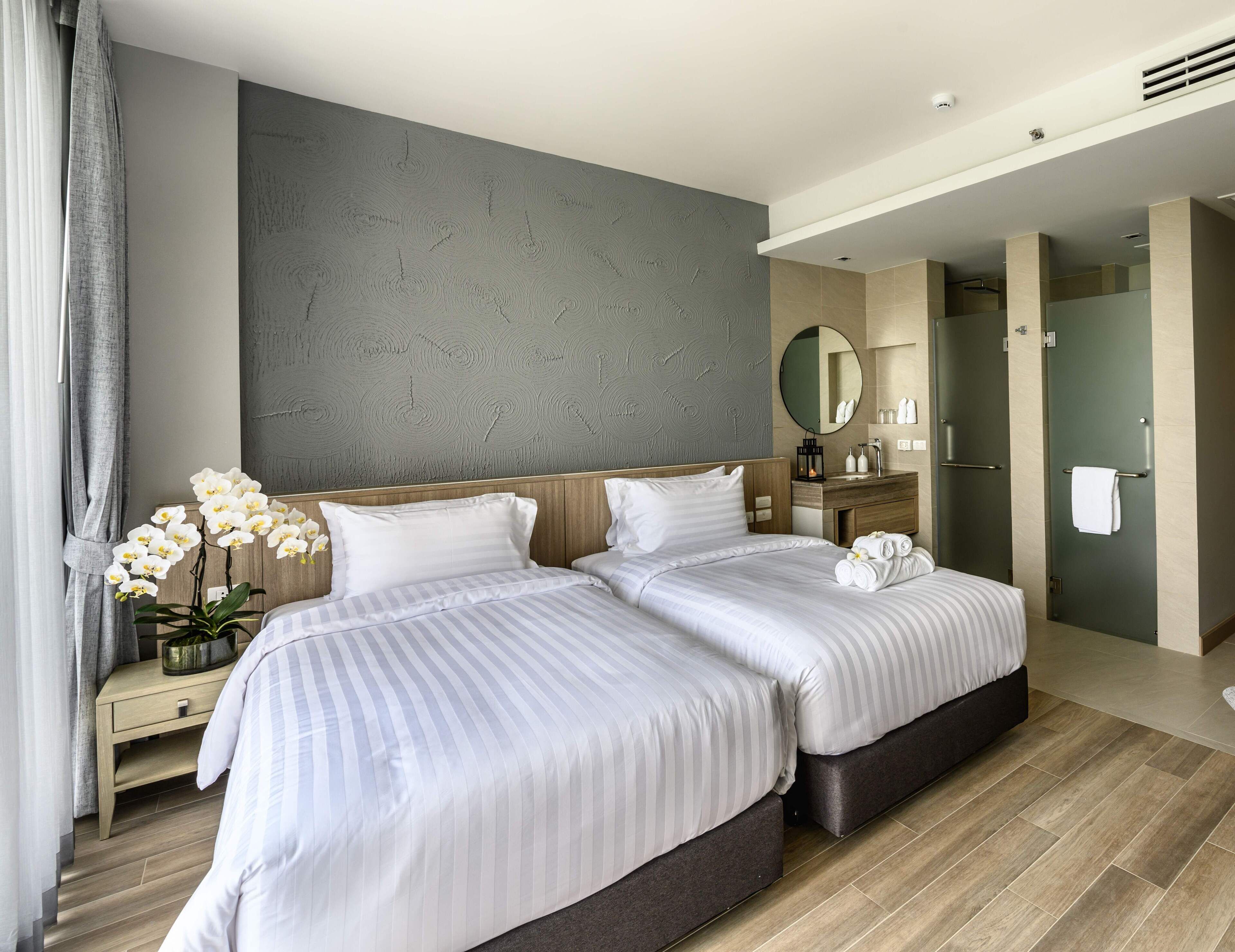 Une chambre d’hôtel avec deux lits, une tête de lit en bois, un mur gris à texture, un miroir rond, une coiffeuse en bois et une porte de salle de bain.