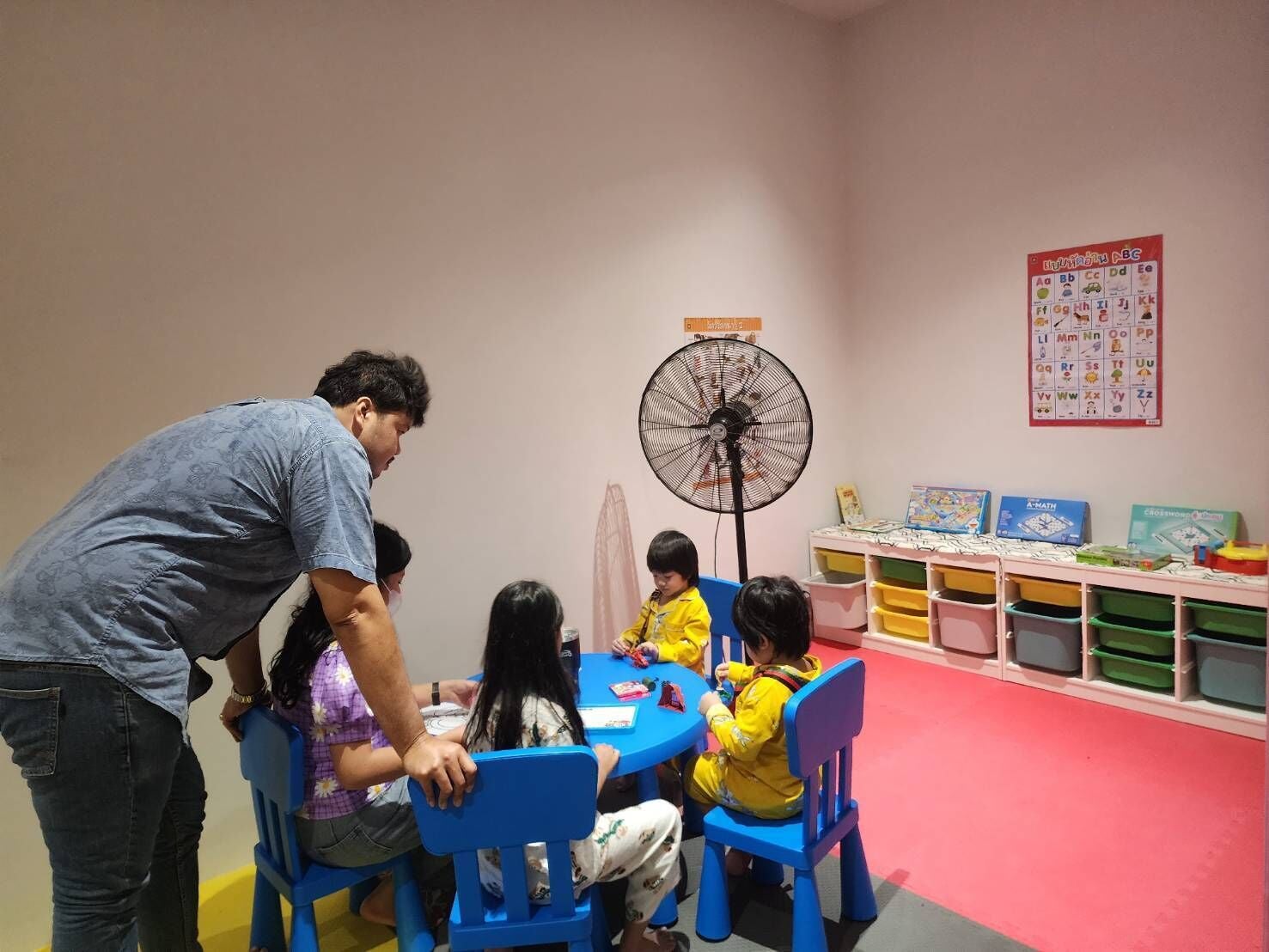 A group of children and an adult at a table with educational materials, in a room with a fan and alphabet posters.