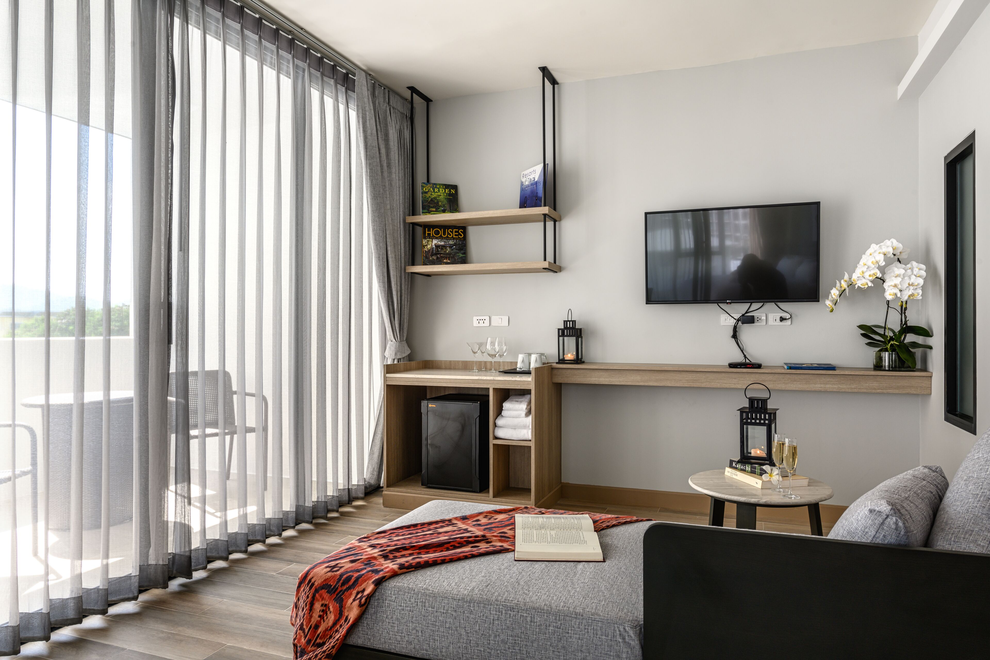 Moderni hotellihuone, jossa on suuri ikkuna, litteänäyttöinen televisio, hyllyillä varustettu työpöytä, pieni pyöreä pöytä, jonka päällä on kirja ja lasit, sekä sohva.