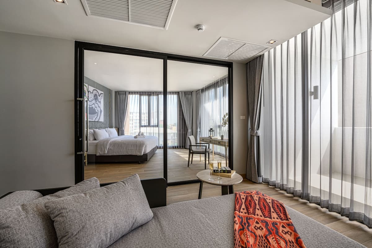 Una habitación de hotel moderna con una cama grande, un sofá, una mesita con un libro y vasos, y un balcón con vistas.