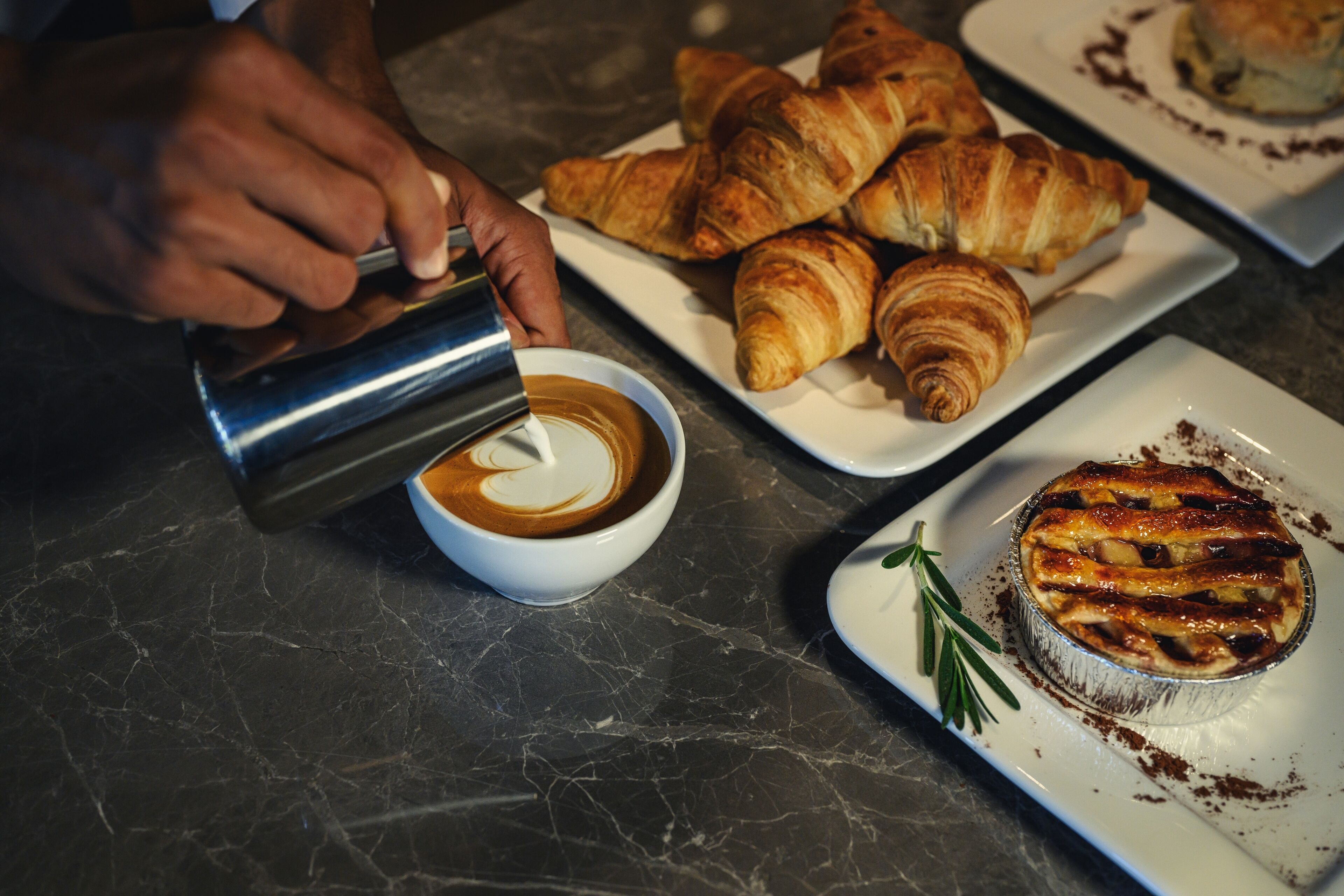 Una persona sirviendo café en una taza, un plato de cruasanes y un pastel horneado sobre una encimera de mármol.