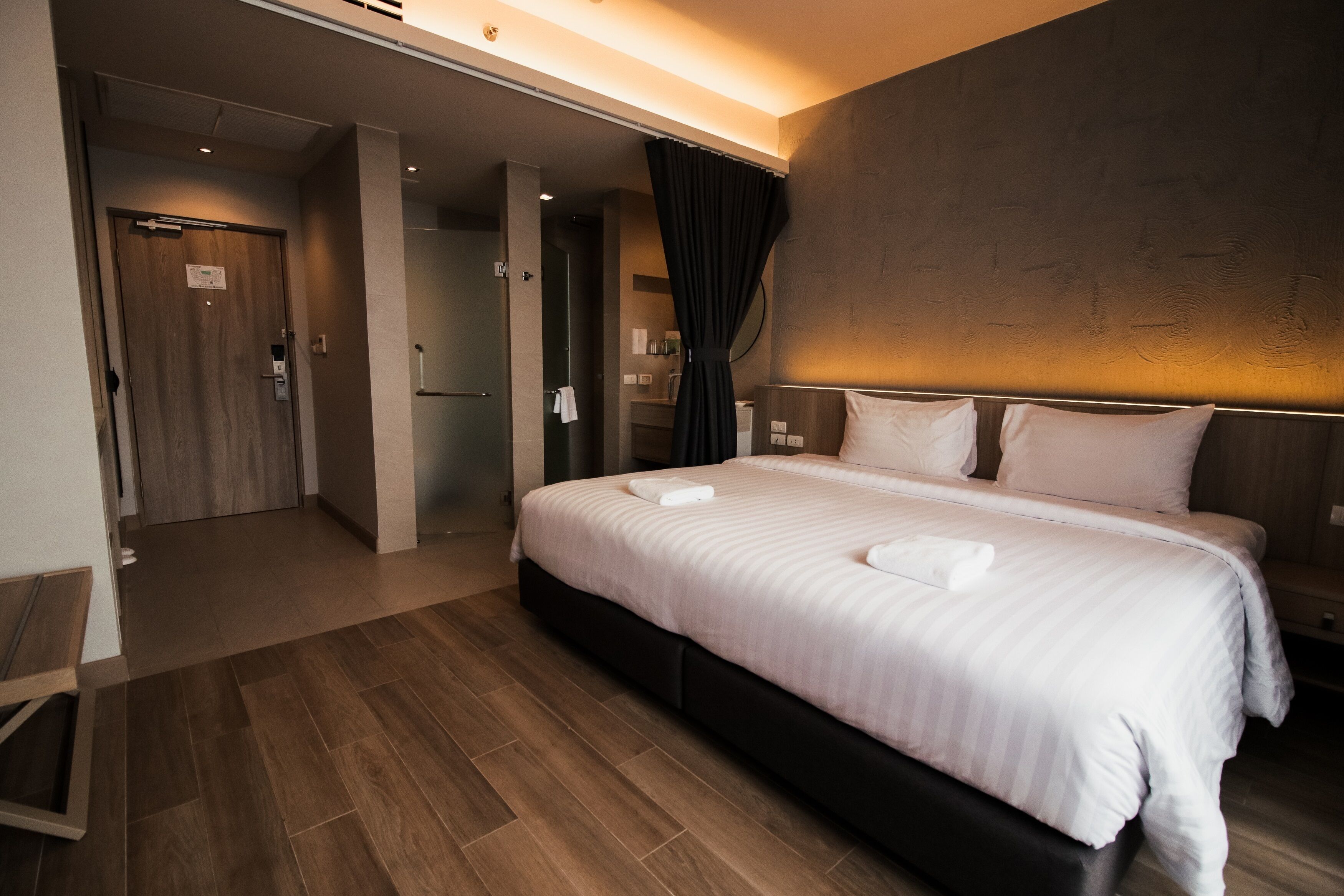 A hotel room with a large bed, a wooden floor, a bathroom with a shower, and a dark curtain.