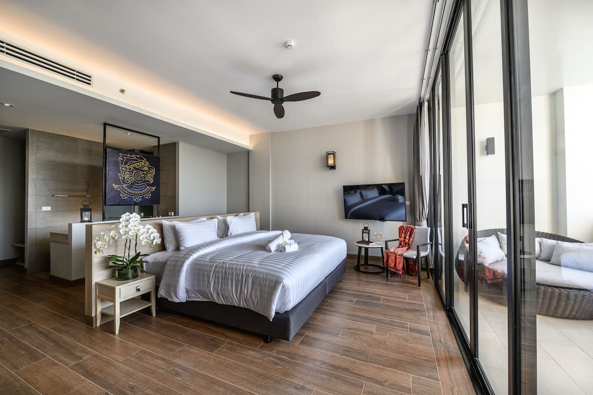 Una habitación de hotel moderna con una cama grande, televisor, zona de estar con una silla y sofá, y un ventanal amplio.