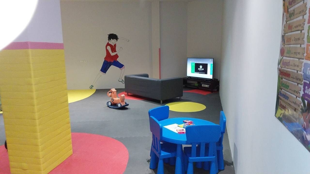 A children's play area with a large yellow pillar, blue chairs, a grey sofa, and a television on a stand.