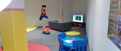 Zona de juegos infantiles con un pilar amarillo grande, sillas azules, un sofá gris y un televisor sobre un mueble.