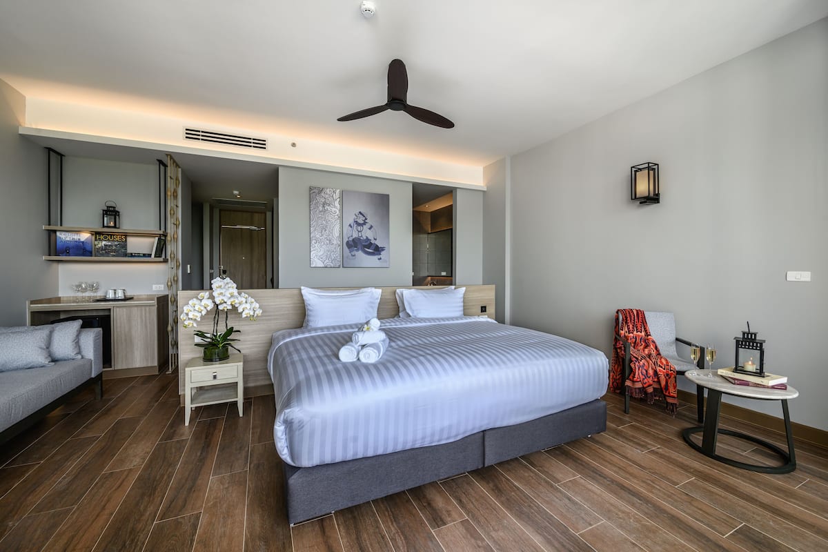 Habitación de hotel moderna con una cama grande, un sofá, una mesita pequeña y una silla. Cuenta con un ventilador de techo, una lámpara de pared e imagen enmarcada.