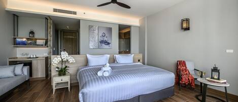 A modern hotel room with a large bed, a sofa, a small table, and a chair. There is a ceiling fan, a wall-mounted light, and a framed picture on the wall.