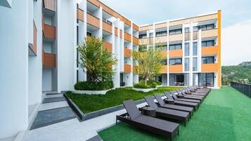A modern building with multiple balconies, a rooftop garden with artificial grass and sun loungers, and a view of a hilly landscape.