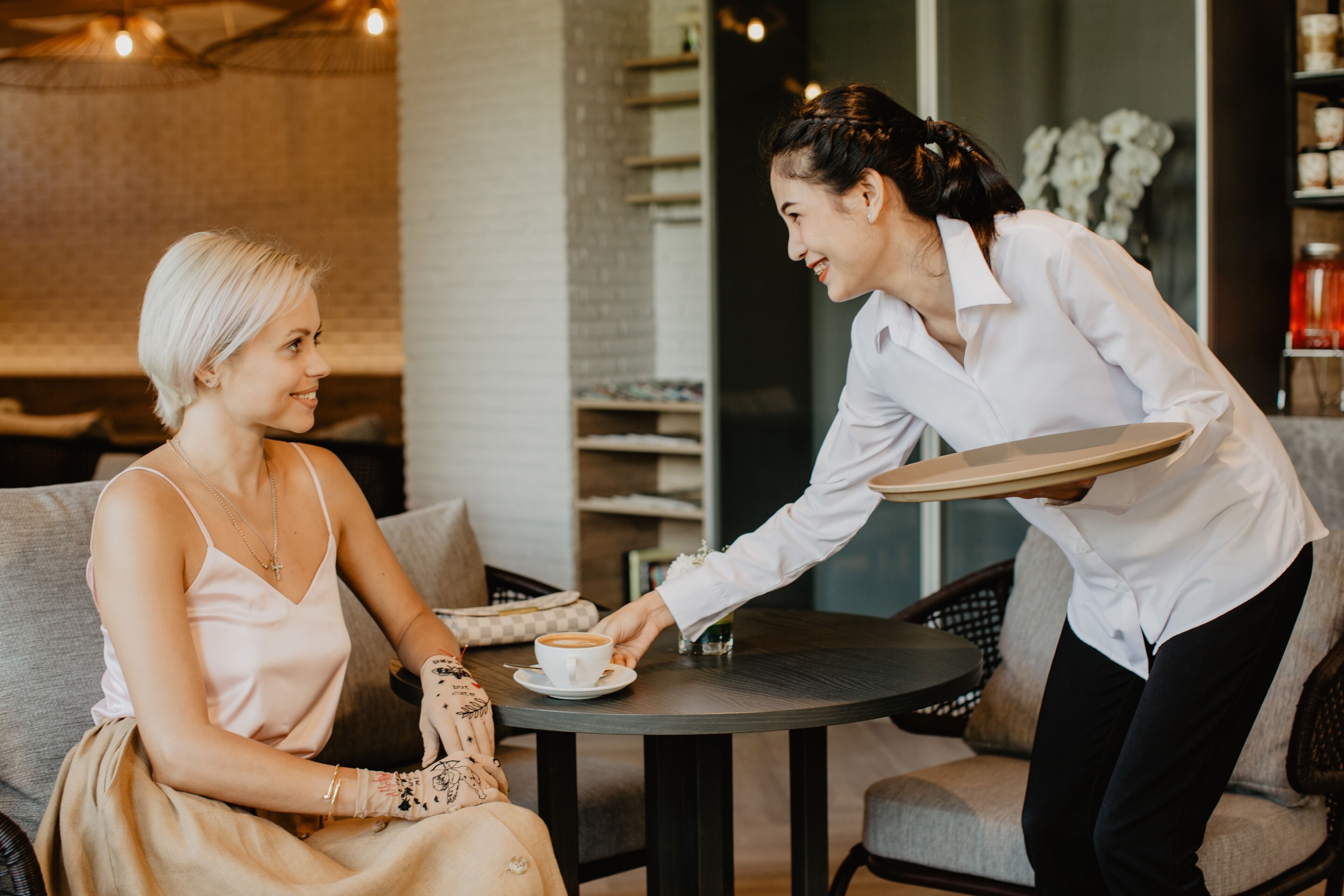 Una mujer sentada en una mesa de café, recibiendo una bandeja con una taza de café.