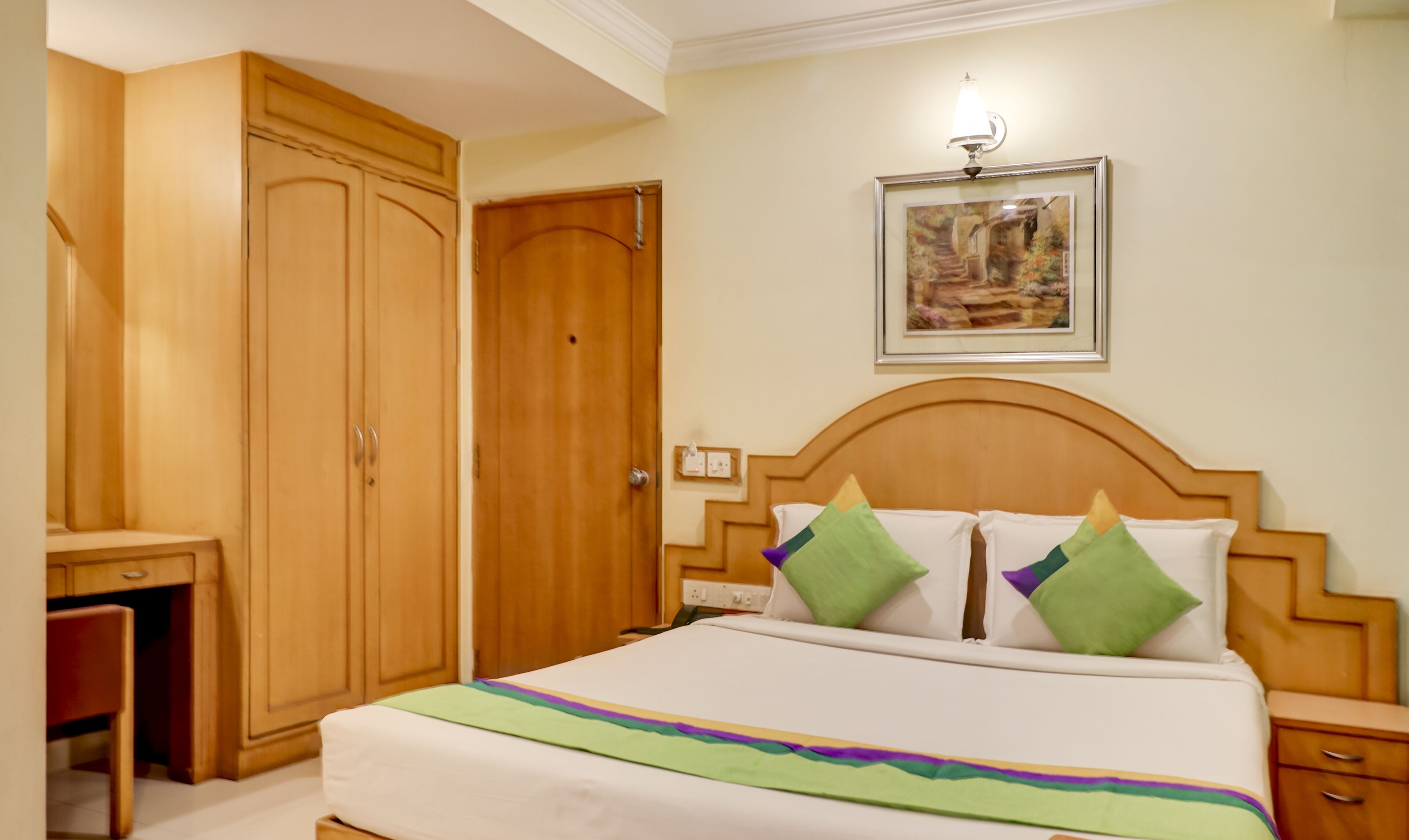 A hotel room with a large wooden wardrobe, a bed with green and white bedding, a nightstand, and a framed picture on the wall.