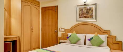 A hotel room with a large wooden wardrobe, a bed with green and white bedding, a nightstand, and a framed picture on the wall.