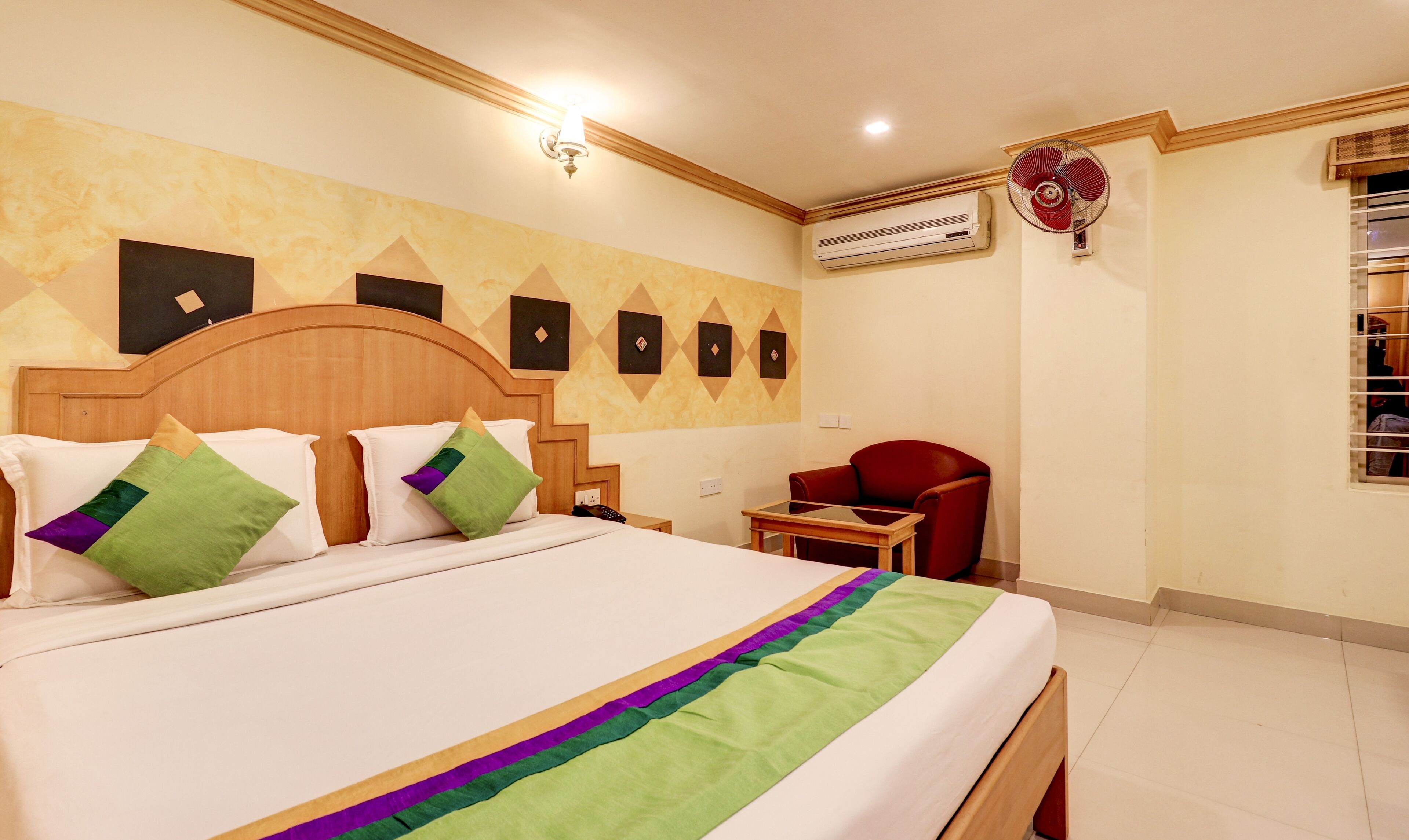 Quarto de hotel com cama de madeira, uma cadeira vermelha, ar condicionado e parede com padrões geométricos.