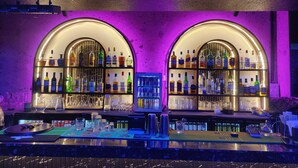 A bar with two illuminated shelves of liquor bottles, a neon sign above stating "THIS IS WHERE THE MAGIC BEGINS", and a bar counter with various bar tools and a tablet. - Treebo Terminus, Koramangala 5th Block (Bengaluru)