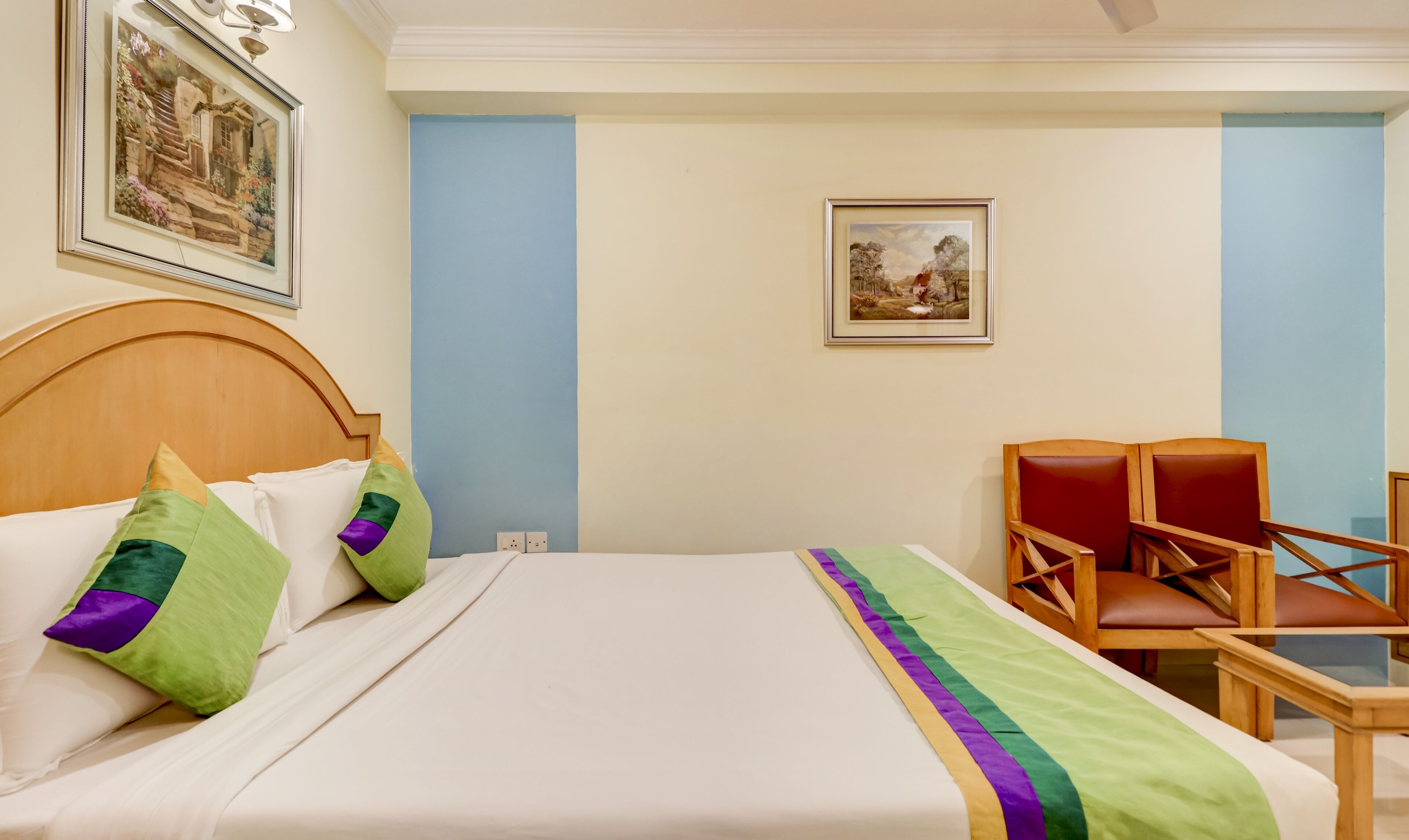 Quarto de hotel com cama, duas almofadas, uma cadeira e duas imagens emolduradas na parede.