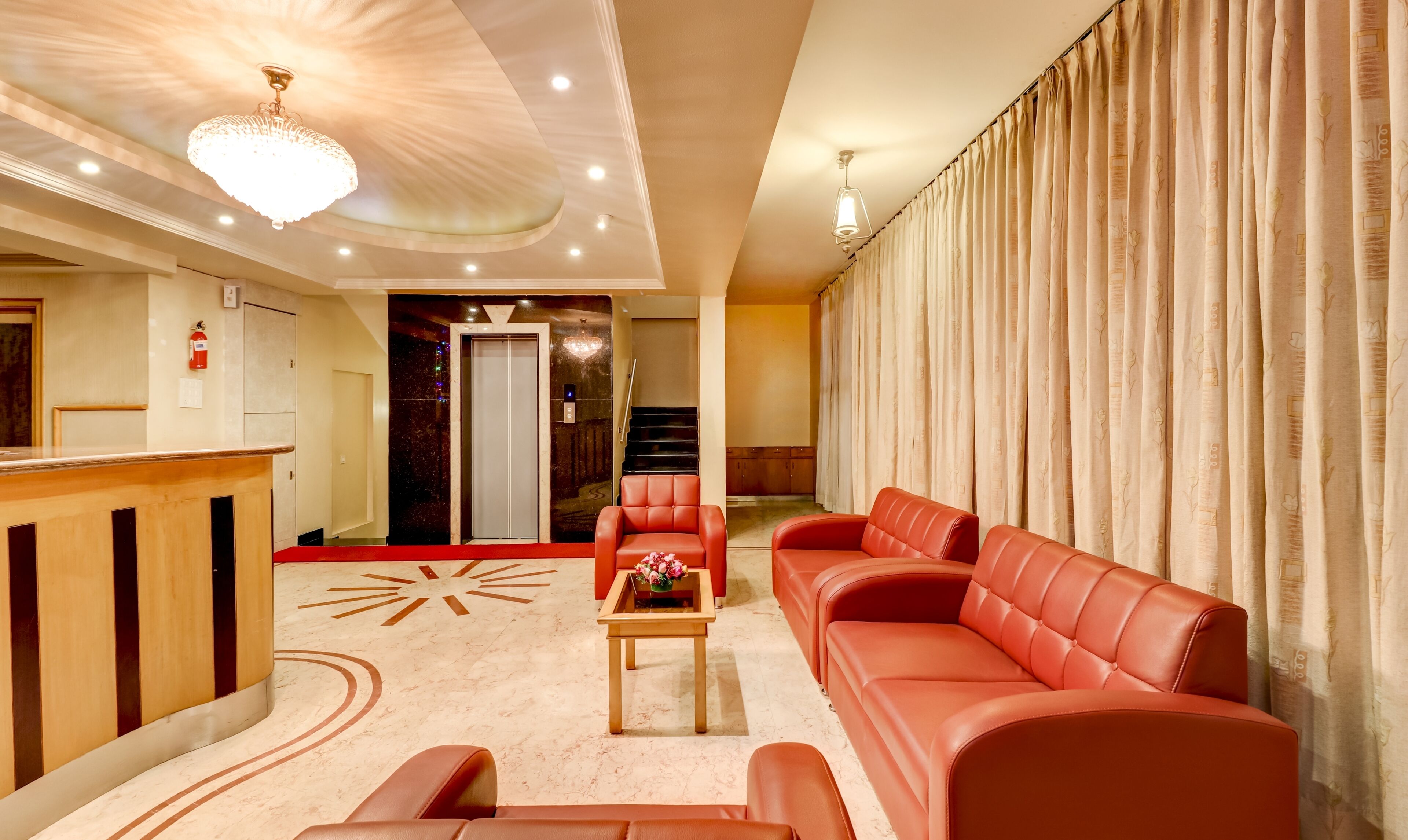 Un'accogliente hall d'albergo con un divano in pelle rossa, una reception in legno, un pavimento in marmo con motivo a raggiera e una scala che conduce a un piano superiore.