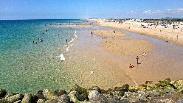 Uma praia com água cristalina, pedras em primeiro plano e pessoas na areia.