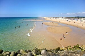 Ein Strand mit klarem Wasser, Felsen im Vordergrund und Menschen im Sand.