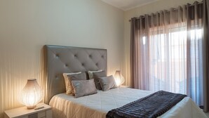 Cama bem arrumada com cabeceira estofada, abajur e janela com cortinas.
