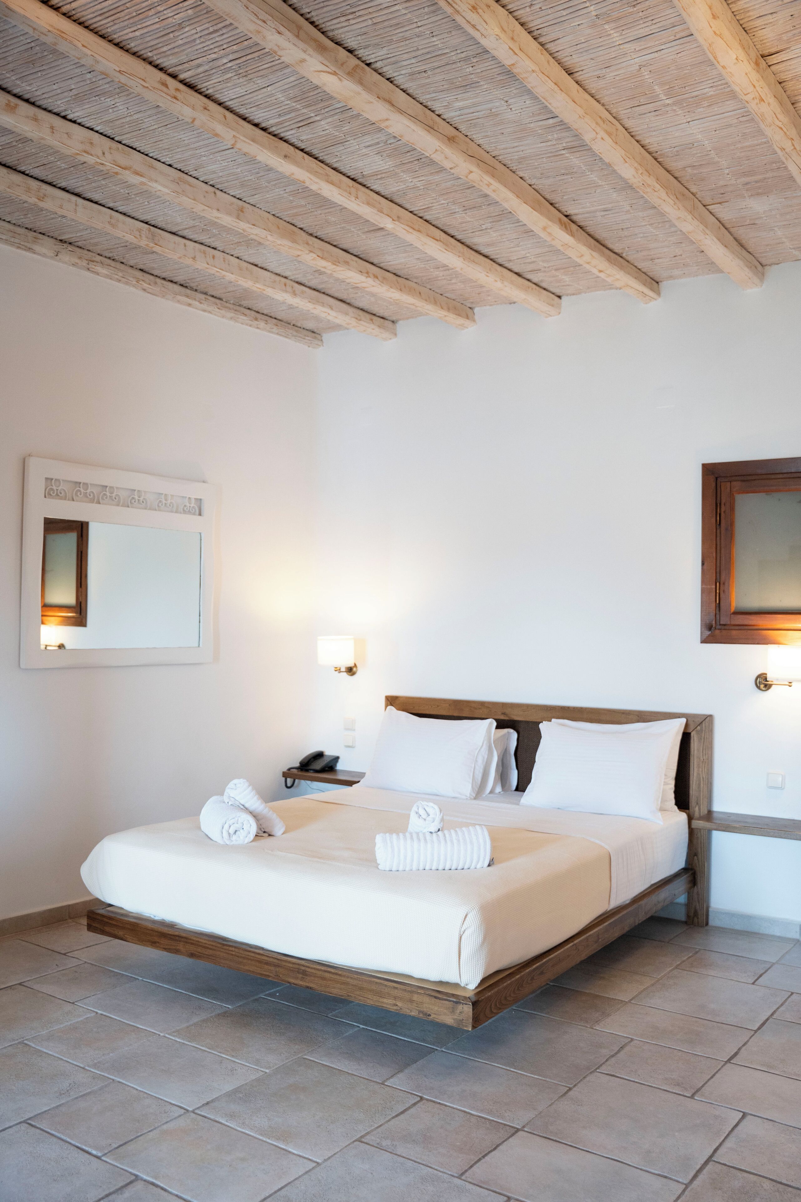 A bedroom with a wooden bed frame, white linens, a bedside table with a lamp, and a mirror on the wall.
