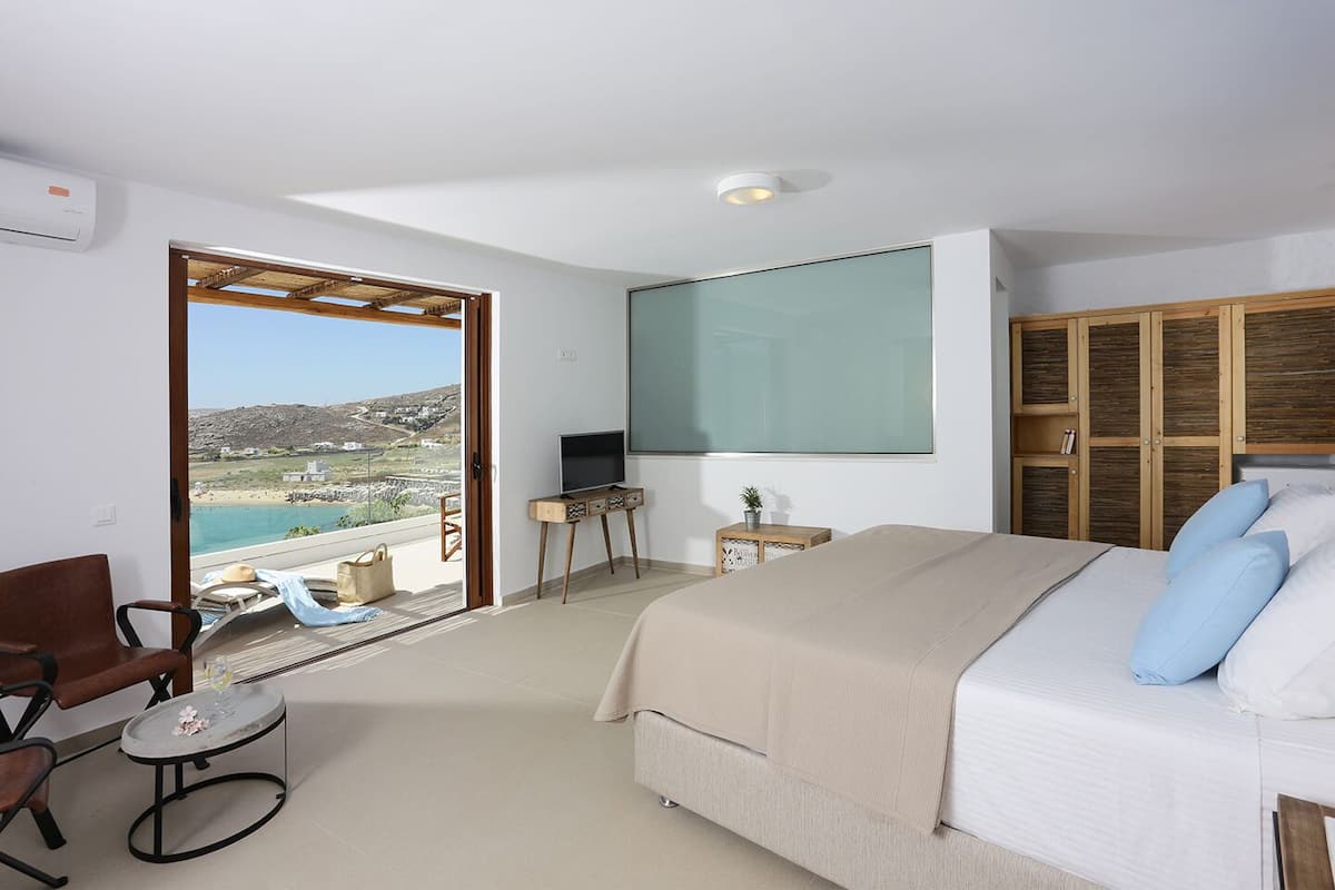 A bedroom with a bed, a TV, a chair, a small table, and a view of the sea.
