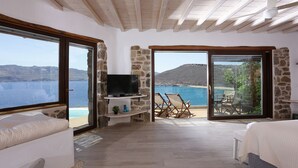 A bedroom with a large window, a TV on a stone shelf, and a balcony with a view of the sea. - Albatros Club Mykonos (Mykonos)