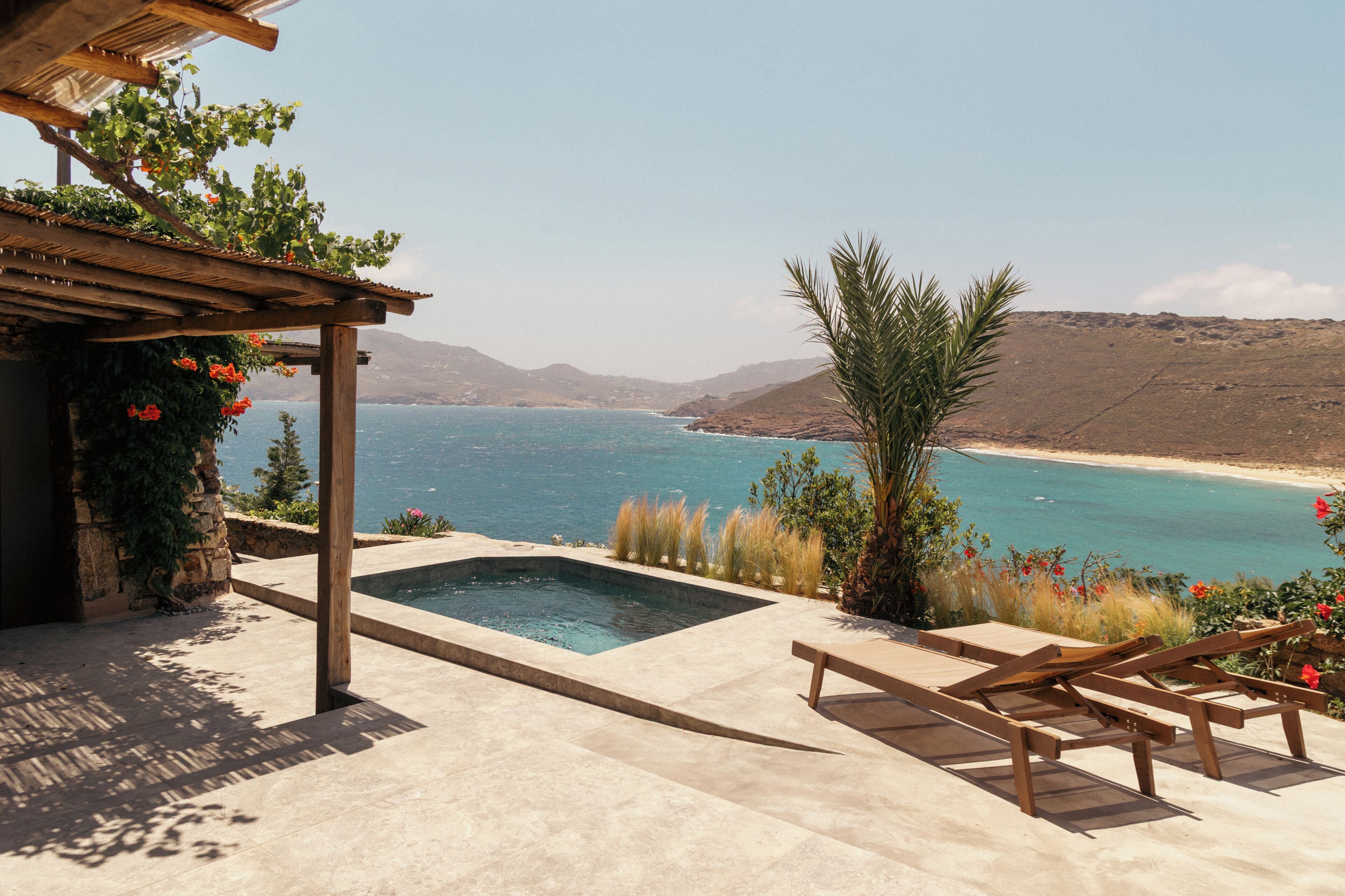 A pool with a view of the sea, wooden lounge chairs, and a pergola with plants.