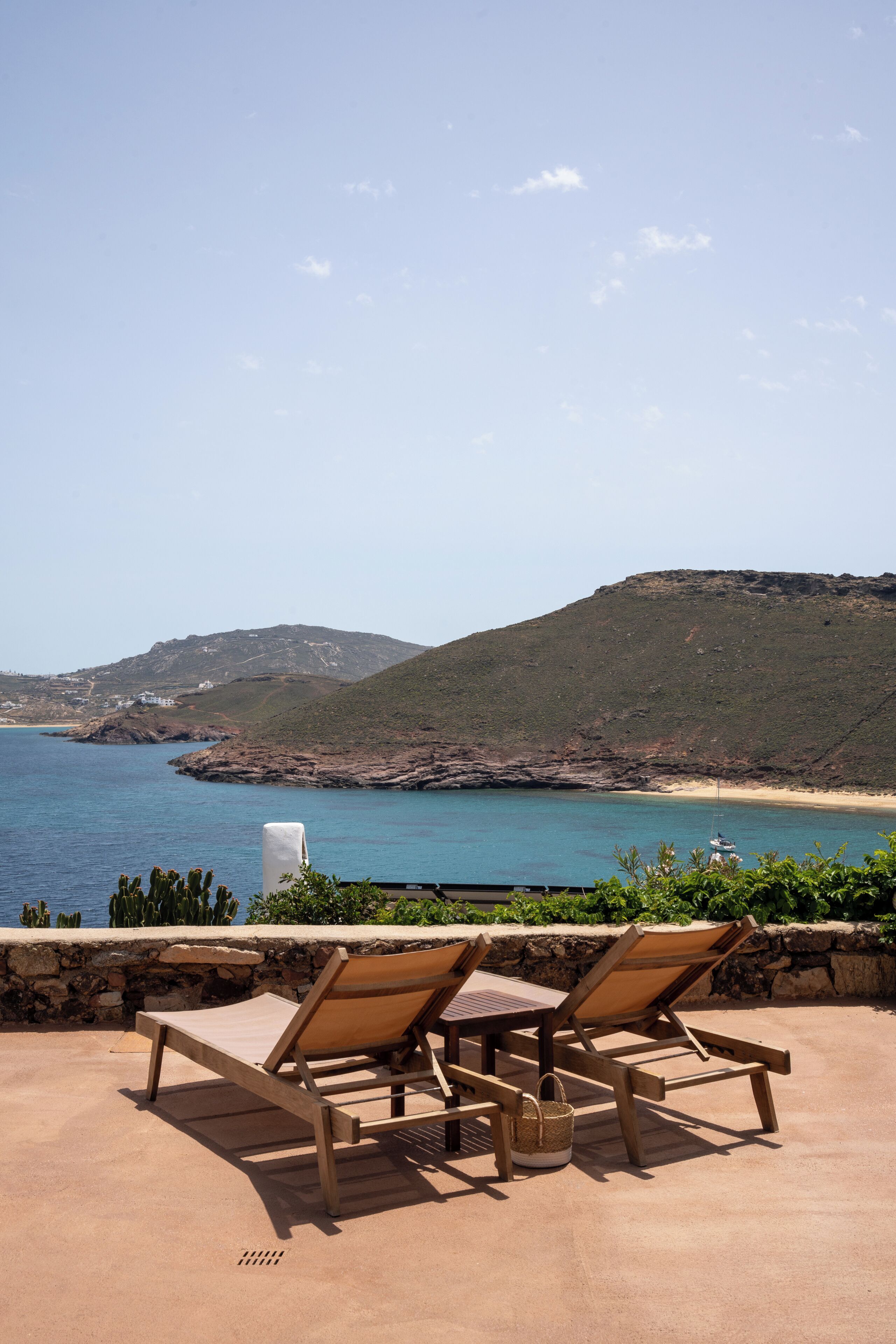 A patio with two wooden lounge chairs, a table, and a stone wall overlooking a beach and sea.