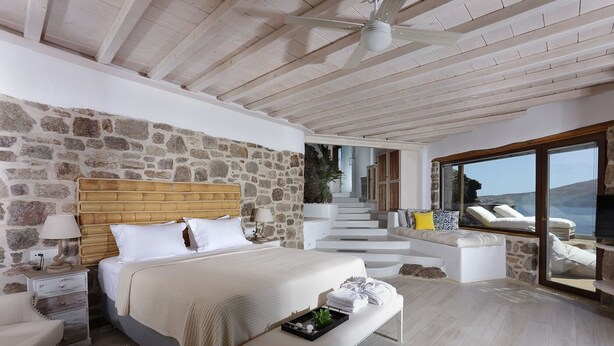 A bedroom with a stone wall, a wooden headboard, a bed with white linens, a bench, a nightstand, and a view of the sea through large windows. - Albatros Club Mykonos (Mykonos)