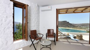 A room with a white textured wall, a wooden door, two brown leather chairs, a small round table with a glass, and a view of a beach and mountains through a glass door. - Albatros Club Mykonos (Mykonos)