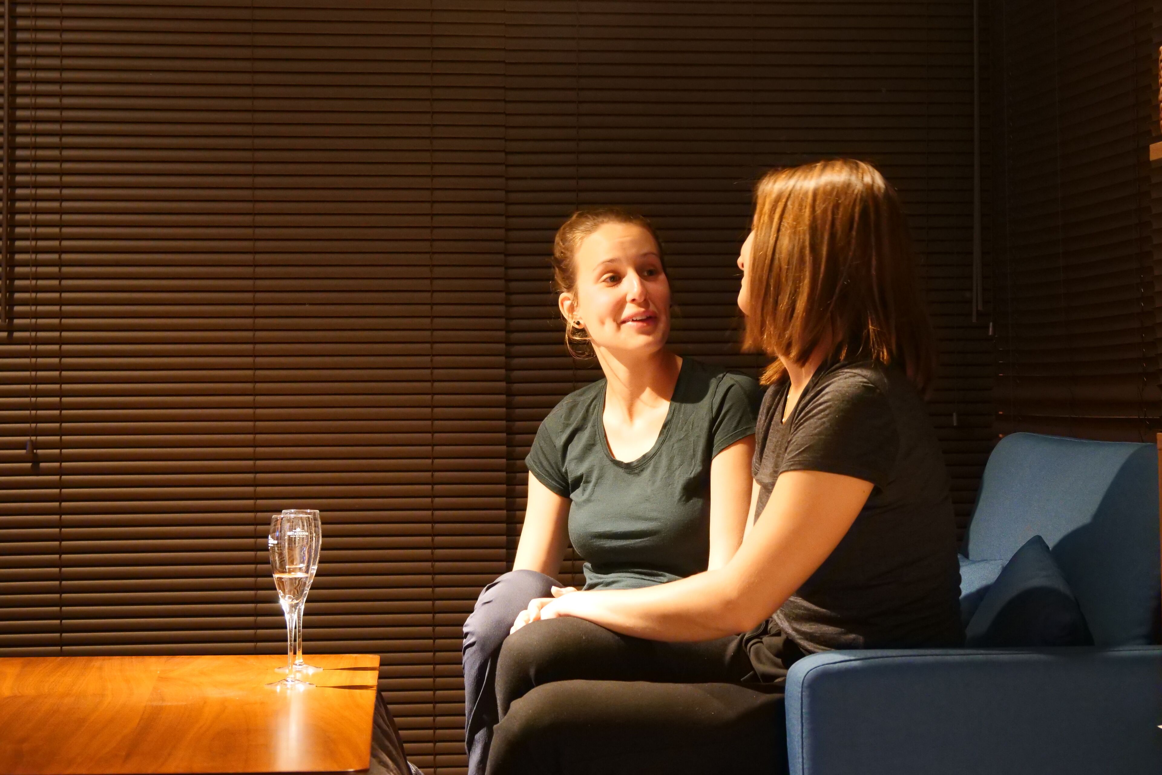 Two people sitting on a blue couch with a wooden table and a glass of water in front of them.