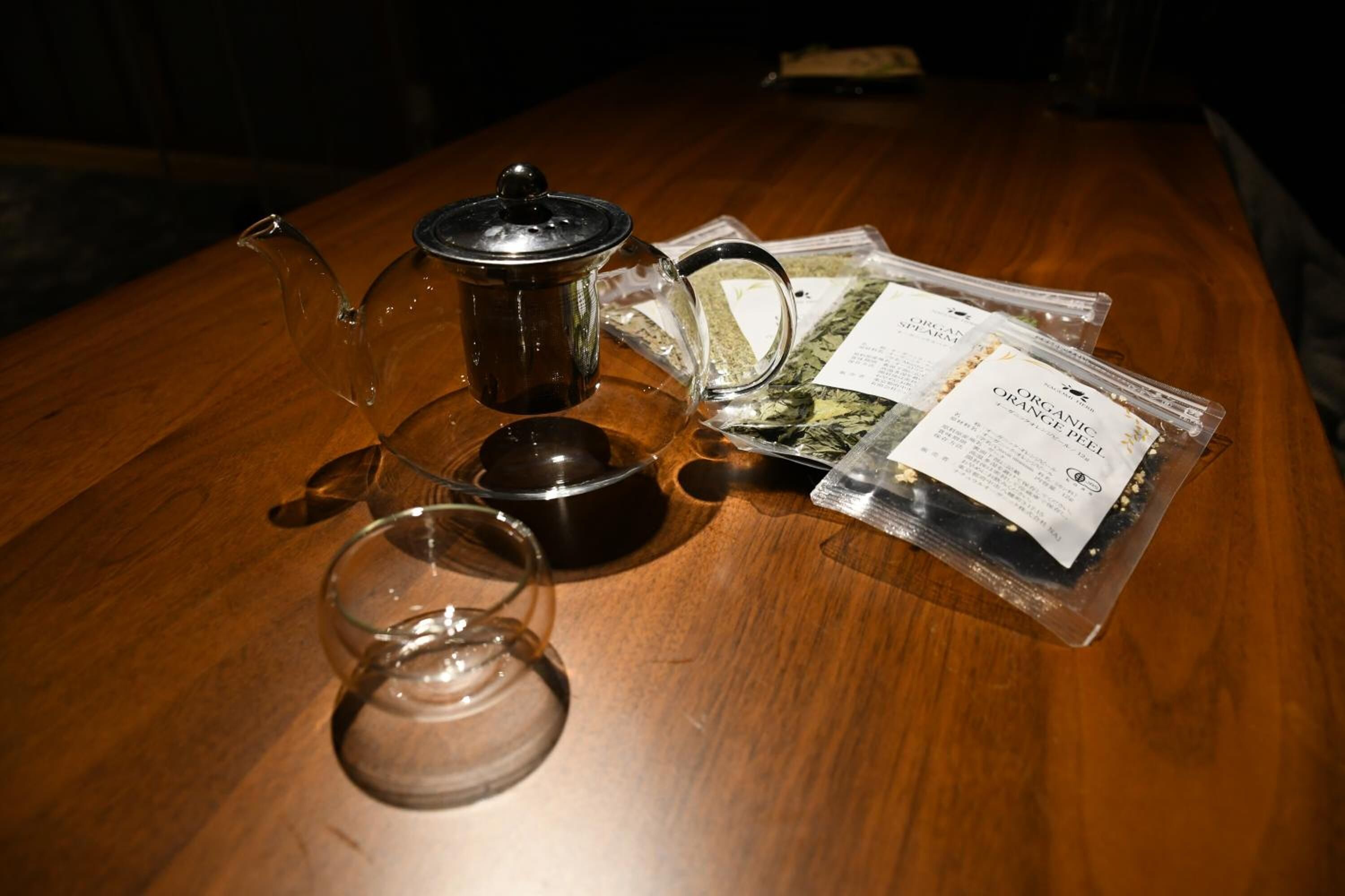 A wooden table with a glass teapot, a glass cup, a black container, and packets of organic orange peel tea.